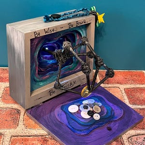 May include: A mixed-media art piece featuring a gray wooden box with the words "Be Wise... Be Brave... Be Tricky." Inside the box is a layered, colorful abstract design. A wire sculpture extends from the box, and buttons are arranged on a purple base.