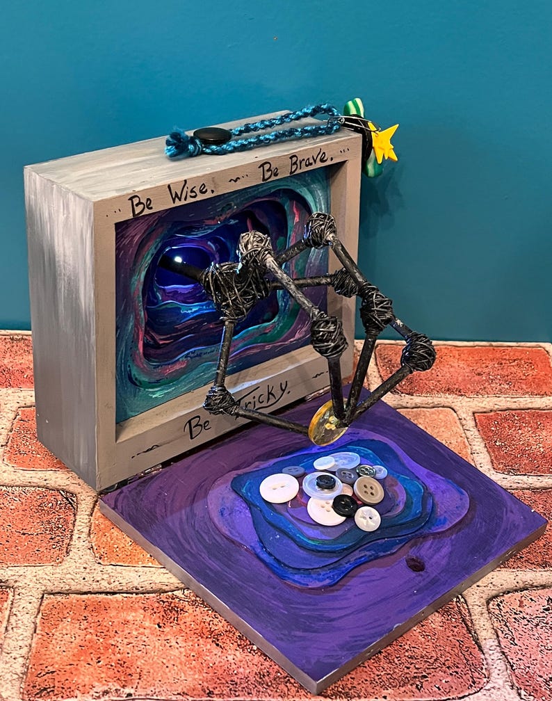 May include: A mixed-media art piece featuring a gray wooden box with the words "Be Wise... Be Brave... Be Tricky." Inside the box is a layered, colorful abstract design. A wire sculpture extends from the box, and buttons are arranged on a purple base.