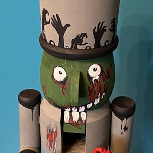 May include: A hand-painted wooden nutcracker in the form of a zombie. The zombie has a green face with white teeth, blood dripping from its eyes, and a gray top hat with black zombie hands. It holds a knife and a red rose.