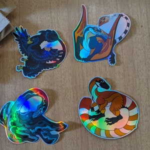 Pu&ograve; includere: Quattro adesivi olografici iridescenti con diverse creature mitiche. Gli adesivi sono su una superficie di legno. Le creature sono un drago blu e arancione, un serpente verde e blu, un dinosauro marrone e arancione e un uccello nero e blu.