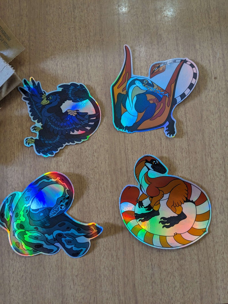 Pu&ograve; includere: Quattro adesivi olografici iridescenti con diverse creature mitiche. Gli adesivi sono su una superficie di legno. Le creature sono un drago blu e arancione, un serpente verde e blu, un dinosauro marrone e arancione e un uccello nero e blu.