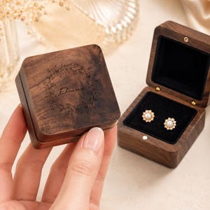 May include: A square dark wood jewelry box with a hinged lid featuring a laser-engraved floral wreath and the name Eliana. The open box interior is lined with black velvet and contains a pair of gold-toned stud earrings with central pearls.