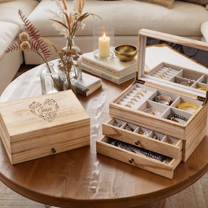 May include: Two wooden jewelry boxes on a round, brown table. One box is closed with the word "Grace" and floral design. The other box is open, revealing multiple compartments and drawers filled with jewelry. A candle and vase of flowers are also on the table.