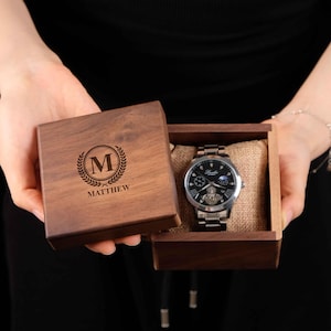 Puede incluir: Un reloj de pulsera plateado con esfera negra se muestra en una caja de madera abierta. La caja está grabada con un monograma y el nombre "MATTHEW". El reloj tiene una correa plateada y se presenta sobre un cojín de yute.