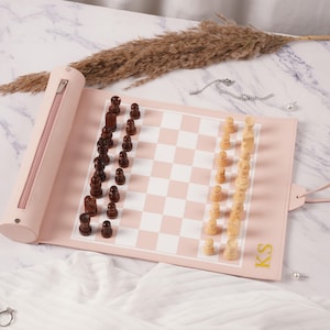 Può includere: Un set di scacchi arrotolabile con un tappetino in similpelle rosa chiaro e una scacchiera a riquadri bianchi e rosa. Il set include pezzi degli scacchi in legno marrone scuro da un lato e pezzi in legno naturale chiaro dall'altro. Le lettere KS sono ricamate in oro.