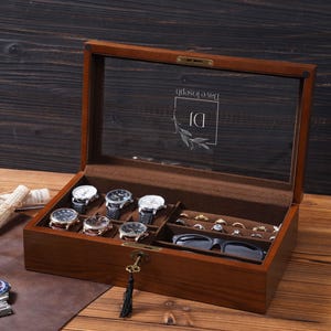 May include: A wooden watch and jewelry box with a glass top. The box contains several watches, rings, and a pair of glasses. The glass top has the text "Dave Joseph" and "DJ" with a floral design. The box is locked with a key.
