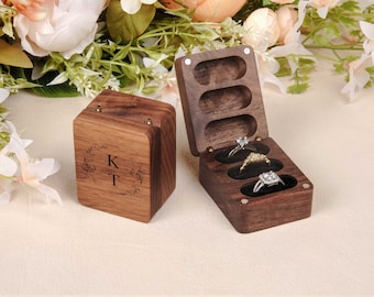 May include: A wooden ring box with three compartments, each with a velvet lining. The box is open and has three rings inside. The box is engraved with the initials "K" and "T" and a floral design.
