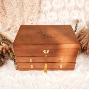 May include: A rectangular wooden jewelry box with a medium brown finish, featuring two drawers with small gold knobs and a top compartment with a gold lock and tassel. The lid is engraved with a floral line art illustration and the name Alex.