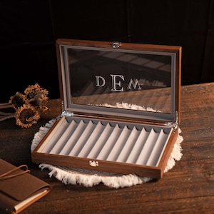 Może przedstawiać: Otwarta drewniana gablota na długopisy ze szklanym wieczkiem. Wnętrze wyłożone jest szarą aksamitną wyści&oacute;łką z indywidualnymi przegr&oacute;dkami na długopisy. Inicjały "DEM" są wytrawione na szkle.