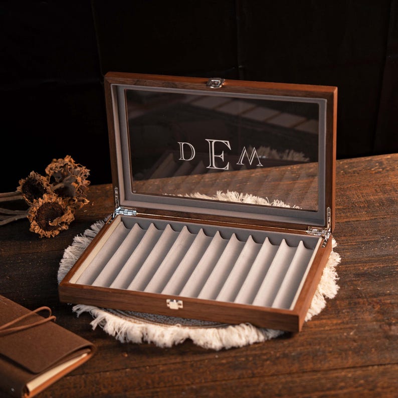 Może przedstawiać: Otwarta drewniana gablota na długopisy ze szklanym wieczkiem. Wnętrze wyłożone jest szarą aksamitną wyści&oacute;łką z indywidualnymi przegr&oacute;dkami na długopisy. Inicjały "DEM" są wytrawione na szkle.