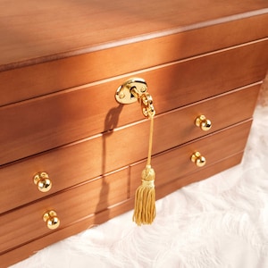 May include: A wooden jewelry box with a smooth, medium-brown finish featuring three drawers with polished gold-tone spherical knobs. A gold-tone key is inserted into the top lock, attached to a matching gold-tone decorative tassel hanging down.