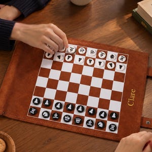 May include: A roll-up brown suede chess board with a white and brown grid. The board features round, flat game pieces with black and white chess icons. The name Clare is embossed in gold on the right side of the suede border.