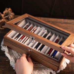 Może przedstawiać: Drewniane etui na długopisy ze szklaną pokrywą, prezentujące kolekcję długopis&oacute;w w r&oacute;żnych kolorach. Etui jest otwarte, ukazując długopisy starannie ułożone w indywidualnych przegr&oacute;dkach. Drewno ma ciepły, naturalny odcień.
