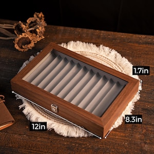 Może przedstawiać: Drewniane etui na długopisy ze szklanym blatem, o długości 30.5 cm, szerokości 21.1 cm i wysokości 4.3 cm. Wnętrze posiada szare, aksamitne przegr&oacute;dki. Etui stoi na dekoracyjnej, frędzlastej macie.