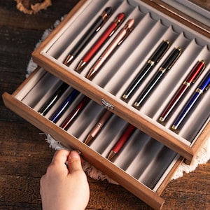 Może przedstawiać: Drewniane etui ekspozycyjne na długopisy z dwoma poziomami, każdy wyłożony szarym filcem i mieszczący r&oacute;żne długopisy. Długopisy są w kolorze czarnym, czerwonym, r&oacute;żowego złota i niebieskim ze złotymi akcentami. Ręka otwiera dolną szufladę.