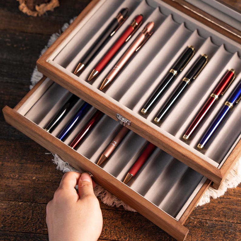 Może przedstawiać: Drewniane etui ekspozycyjne na długopisy z dwoma poziomami, każdy wyłożony szarym filcem i mieszczący r&oacute;żne długopisy. Długopisy są w kolorze czarnym, czerwonym, r&oacute;żowego złota i niebieskim ze złotymi akcentami. Ręka otwiera dolną szufladę.