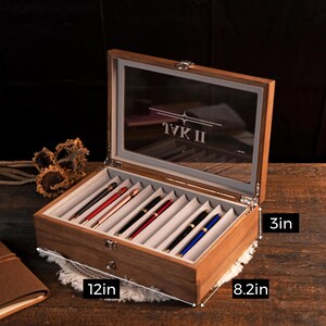 Może przedstawiać: Drewniane etui na długopisy ze szklaną pokrywą, mieszczące kilka długopis&oacute;w w r&oacute;żnych kolorach. Etui ma 30.5 cm długości, 20.8 cm szerokości i 7.6 cm wysokości. Tekst "IVK II" jest widoczny.