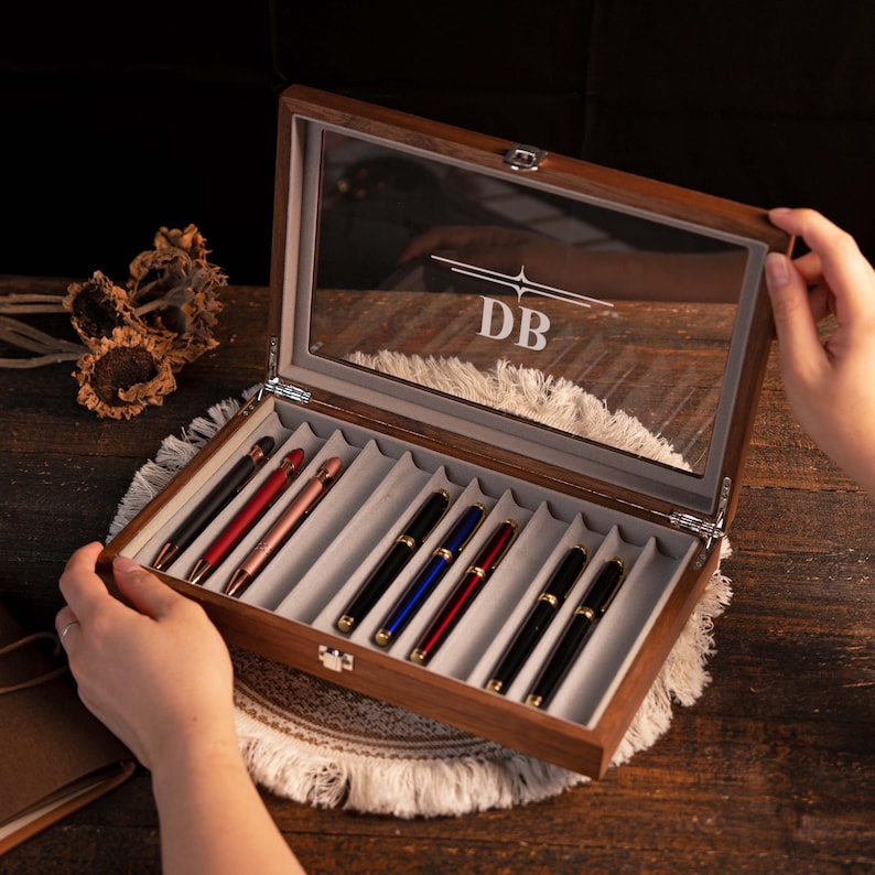 Może przedstawiać: Drewniane etui na długopisy ze szklaną pokrywą, mieszczące kilka długopis&oacute;w w r&oacute;żnych kolorach. Na pokrywie wygrawerowano inicjały "DB". Etui jest otwarte, ukazując długopisy w indywidualnych przegr&oacute;dkach.