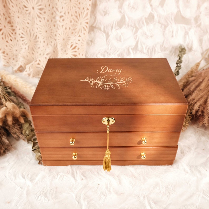 May include: A rectangular wooden jewelry box with a rich brown finish, featuring two pull-out drawers with gold-toned knobs. The lid is engraved with the name Davey, the date 09.11.2025, and a floral branch. A gold key with a tassel hangs from the lock.