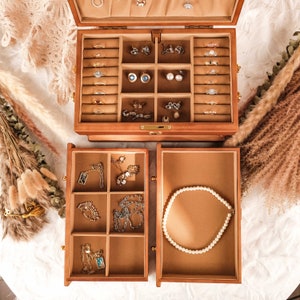 May include: A wooden jewelry box with a hinged lid and two pull-out drawers. The interior is lined with tan fabric and features multiple compartments for rings, earrings, and necklaces. Various gold and silver-toned jewelry pieces are organized inside.