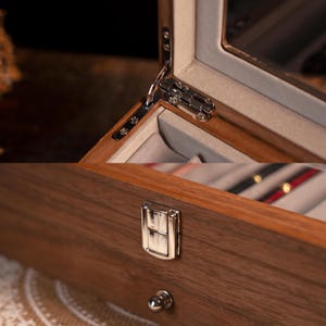 Może przedstawiać: Zbliżenie drewnianego pudełka na biżuterię ze szklanym wieczkiem. Pudełko posiada srebrny zawias i zapięcie. Wnętrze wyłożone jest jasnoszarą tkaniną. Drewno ma bogaty, ciemnobrązowy kolor.