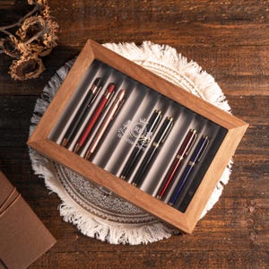 Może przedstawiać: Drewniane etui na długopisy ze szklanym blatem, prezentujące kolekcję długopis&oacute;w w r&oacute;żnych kolorach. Długopisy mają czarne, czerwone i złote akcenty. Etui stoi na dekoracyjnej białej i beżowej podkładce.