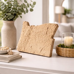 May include: A beige, textured decorative piece in the shape of the state of Oregon. A vase with green foliage, a candle in a glass holder, and a bowl of nuts are also on the white surface.