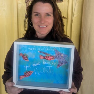 May include: A framed artwork with a white border, featuring a blue background and the words "THERE IS NO GROUND, you must learn to FLOAT." The artwork includes illustrations of red fish and a glittery purple star-shaped figure.