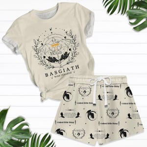 May include: A beige t-shirt and matching shorts set. The t-shirt features a circular graphic with the text "BASGIATH WAR COLLEGE". The shorts have a repeating pattern with the same graphic and text "violent little thing".