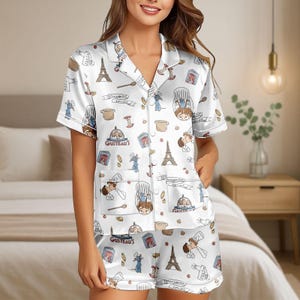 May include: White pyjama set featuring a pattern of cartoon characters, the Eiffel Tower, and cooking motifs. The short-sleeved top has a notched collar and two pockets. The shorts have an elasticated waistband.