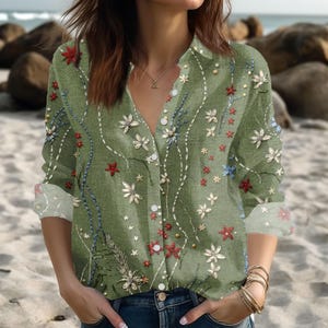 Op de afbeelding: Een groene blouse met knopen en lange mouwen, versierd met een bloemenpatroon in rood, wit en blauw. De blouse heeft een V-hals en is gemaakt van een lichte stof, ideaal voor dagelijks gebruik. Het ontwerp bevat kleine witte stippen.
