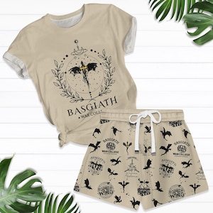 Peut inclure: Ensemble t-shirt et short beiges. Le t-shirt présente un motif noir de dragon et le texte "BASGIATH WAR COLL". Le short a un motif répété de dragons et de texte. Le short a un cordon de serrage blanc.