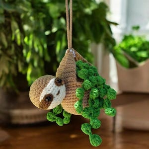 May include: A crocheted sloth hanging from a string with green crocheted leaves. The sloth is brown with a white face and black eyes.