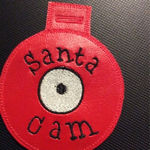 Op de afbeelding: Een rode, cirkelvormige Santa Cam-ornament met zwarte geborduurde tekst en een wit en zwart oogontwerp. Het ornament heeft een kleine lus aan de bovenkant om op te hangen.