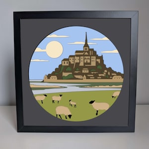 May include: A framed print of a circular illustration depicting Mont Saint-Michel, a historic abbey located on a tidal island in Normandy, France. The illustration features a blue sky with white clouds, a yellow sun, and a green field with sheep.
