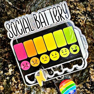 May include: A clear acrylic badge holder with a black and white graphic of a battery with the text "SOCIAL BATTERY" and a rainbow of colored bars. The battery has a lightning bolt and a series of emojis representing different levels of social energy. The badge holder has a rainbow-colored button.
