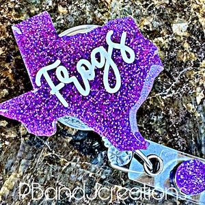 May include: A purple glitter badge reel in the shape of Texas with the word "Frogs" in white script. The badge reel has a clear plastic backing with a metal clip and a purple glitter button.