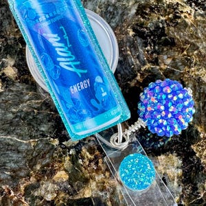 May include: A blue and teal badge reel with a clear plastic strap. The badge reel features a blue Alan Energy drink packet design, a round blue glitter accent, and a blue rhinestone ball.
