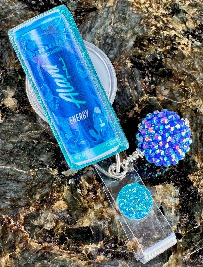 May include: A blue and teal badge reel with a clear plastic strap. The badge reel features a blue Alan Energy drink packet design, a round blue glitter accent, and a blue rhinestone ball.