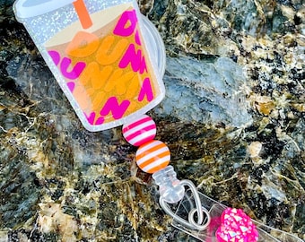 May include: A clear plastic badge reel with a pink and orange striped bead, a pink and white striped bead, and a pink glitter heart charm. The top of the reel features a clear plastic cup with a glittery orange and pink design and a pink straw. The cup has the word "Dunkin'" printed on it.