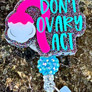 May include: A glittery badge reel with the text "DON'T OVARY ACT" in teal. The design includes a pink ovary and a light blue, gem-like embellishment. The reel has a clear plastic clip and a retractable cord.