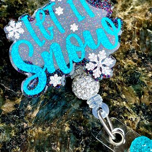 May include: A blue and white glitter badge holder with the text "Let it Snow" and snowflakes. The badge holder has a clear plastic strap and a sparkly blue charm.