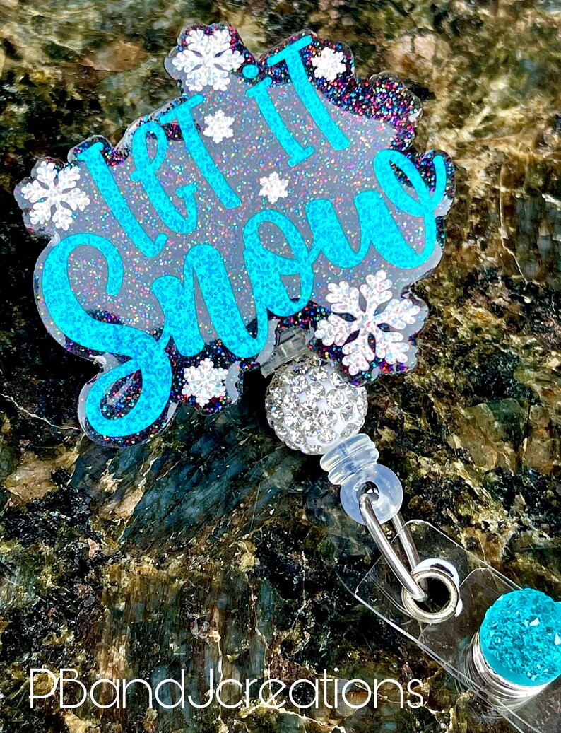 May include: A blue and white glitter badge holder with the text "Let it Snow" and snowflakes. The badge holder has a clear plastic strap and a sparkly blue charm.