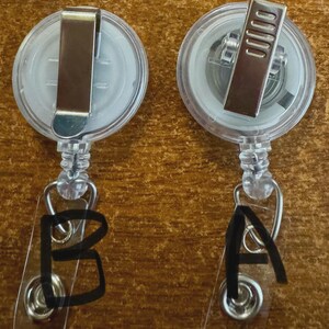 May include: Two clear plastic retractable badge holders with metal clips. One badge holder has the letter "A" written on the clear plastic, and the other has the letter "B". The badge holders are on a wooden surface.
