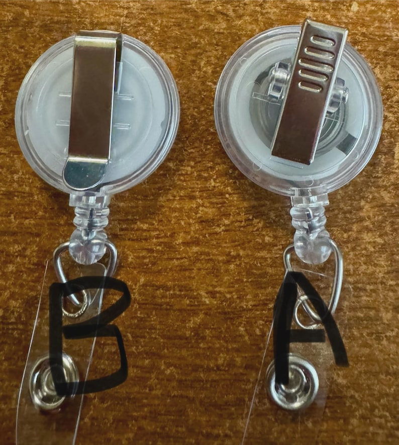May include: Two clear plastic retractable badge holders with metal clips. One badge holder has the letter "A" written on the clear plastic, and the other has the letter "B". The badge holders are on a wooden surface.