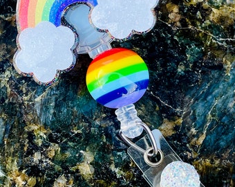 May include: A rainbow-colored retractable badge holder with a rainbow and cloud charm. The charm is made of clear resin with glitter embedded in it. The badge holder has a clear plastic clip and a white glitter circle on the end.