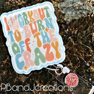 May include: A colorful badge holder with the text "I workout to burn off the crazy" in a retro font. The badge holder has a clear plastic backing and a sparkly pink charm.