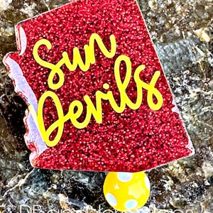 May include: A red glitter badge reel with the words "Sun Devils" in yellow script. The badge reel has a yellow and white polka dot design and a clear plastic backing.
