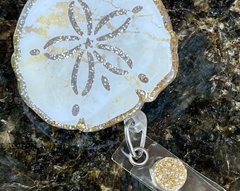 Puede incluir: Un dólar de arena blanco con detalles de purpurina dorada y un diseño de estrella de mar de purpurina plateada. El dólar de arena está unido a un portagafete de plástico transparente con un círculo de purpurina dorada.