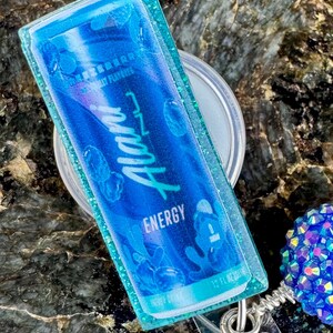 May include: A blue and teal badge reel featuring a miniature replica of an energy drink can. The can design includes the word "Alani" in white script, with "Energy" below. The badge reel has a clear plastic backing and a glittery teal border.